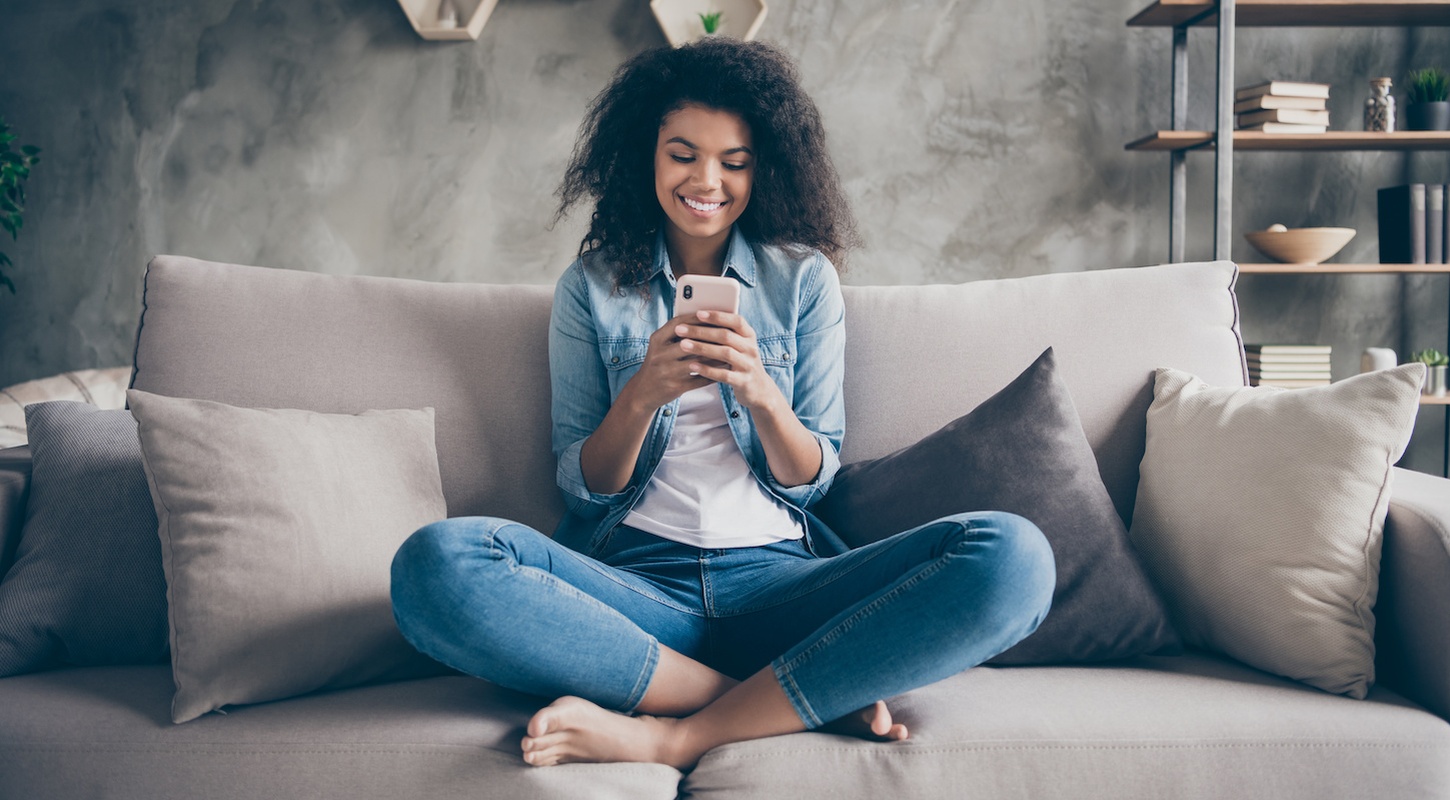 Photo of pretty dark skin curly lady homey domestic texting telephone with friends reading instagram comments sitting comfy couch casual denim outfit living room indoors